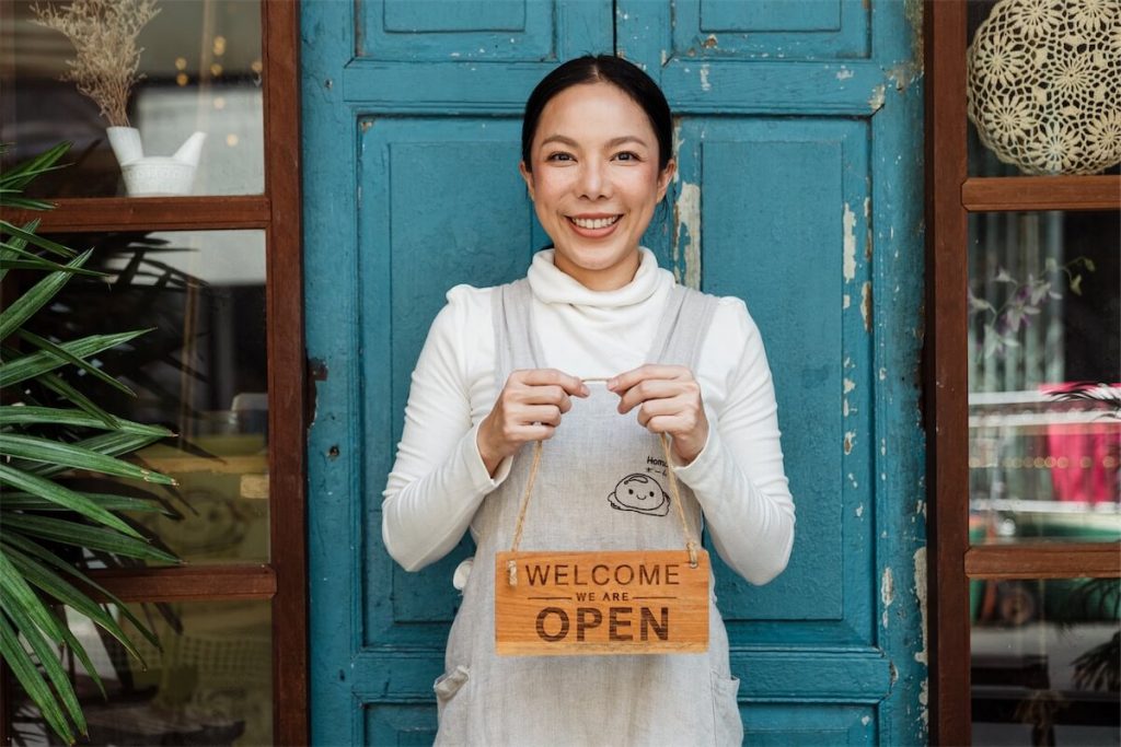 Woman holding a ‘Welcome, we are open’ sign in front of her shop, where some financial institution will only offer business loans for businesses in operation for at least 6 months