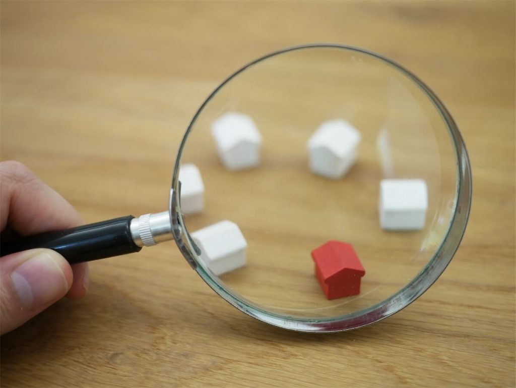 A magnifying glass looking at miniature housing. One of them coloured in red to illustrate rising property prices in Singapore due to rising interest rates.